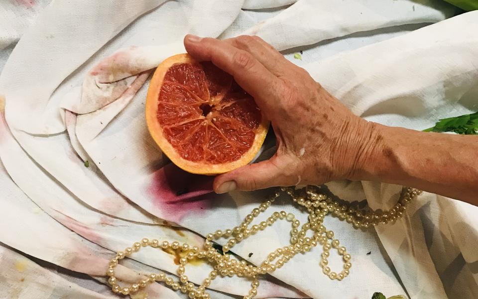 Auf einem bekleckerten Tischtuch. Eine faltige Hand hält eine aufgeschnittene Blutorange. Um sie herum Essensreste und eine Goldkette.