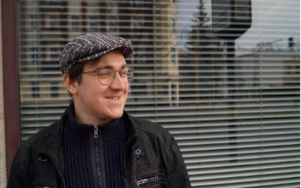 Porträt von Marius Zoschke mit schwarzer Jacke und Schiebermütze, hinter ihm ein Fenster, darin spiegelt sich die Stadt