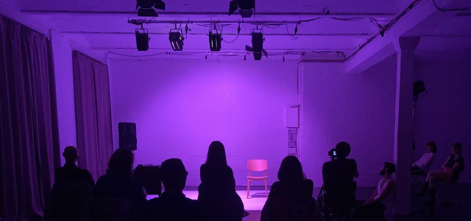 TATWERK Aufnahme einer in Violett beleuchteten Bühne. Die Bühne, die hinter dem Publikum zu sehen ist, ist leer.