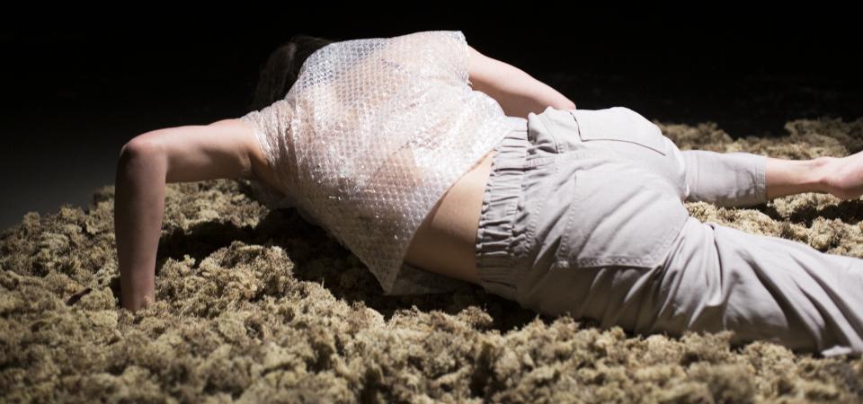 Eine Person liegt auf einer hellen Fläche aus organischem Material - Moos. Die Person ist von abgwandt, stützt sich mit den Händen etwas hoch und hält den Kopf gesenkt. Die Person trägt eine helle Hose und ein kurzärmliges Shirt aus Luftpolsterfolie. Der Hintergrund ist schwarz. 
