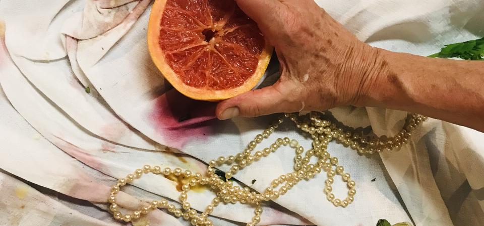 Auf einem bekleckerten Tischtuch. Eine faltige Hand hält eine aufgeschnittene Blutorange. Um sie herum Essensreste und eine Goldkette. 