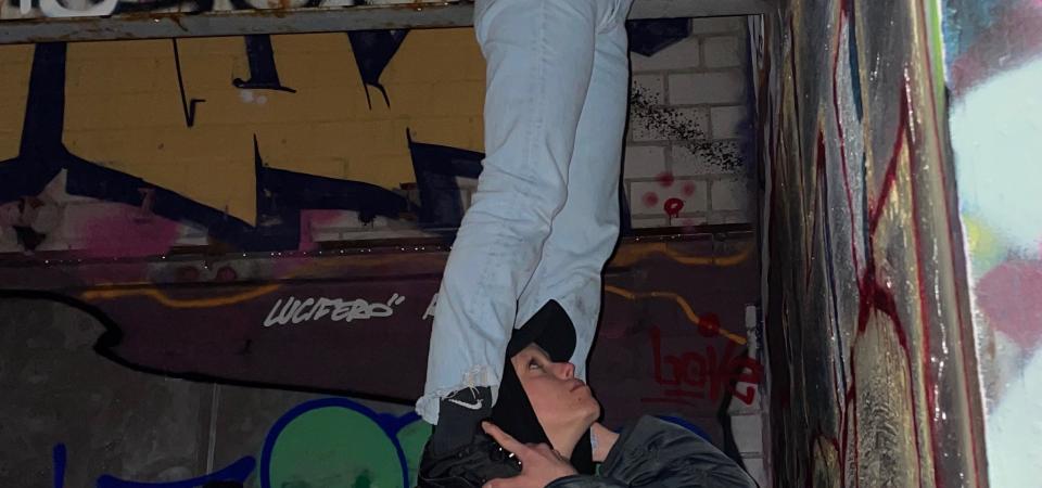 In front of a wall with graffiti, a person stands on the shoulders of another person and reaches up to the ceiling. The person below is also looking up.